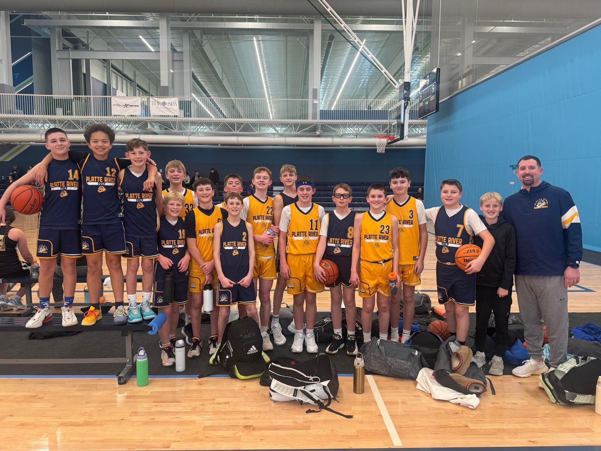 Youth basketball team posing with coaches in a gym, wearing navy and yellow uniforms with trophies and basketballs.
