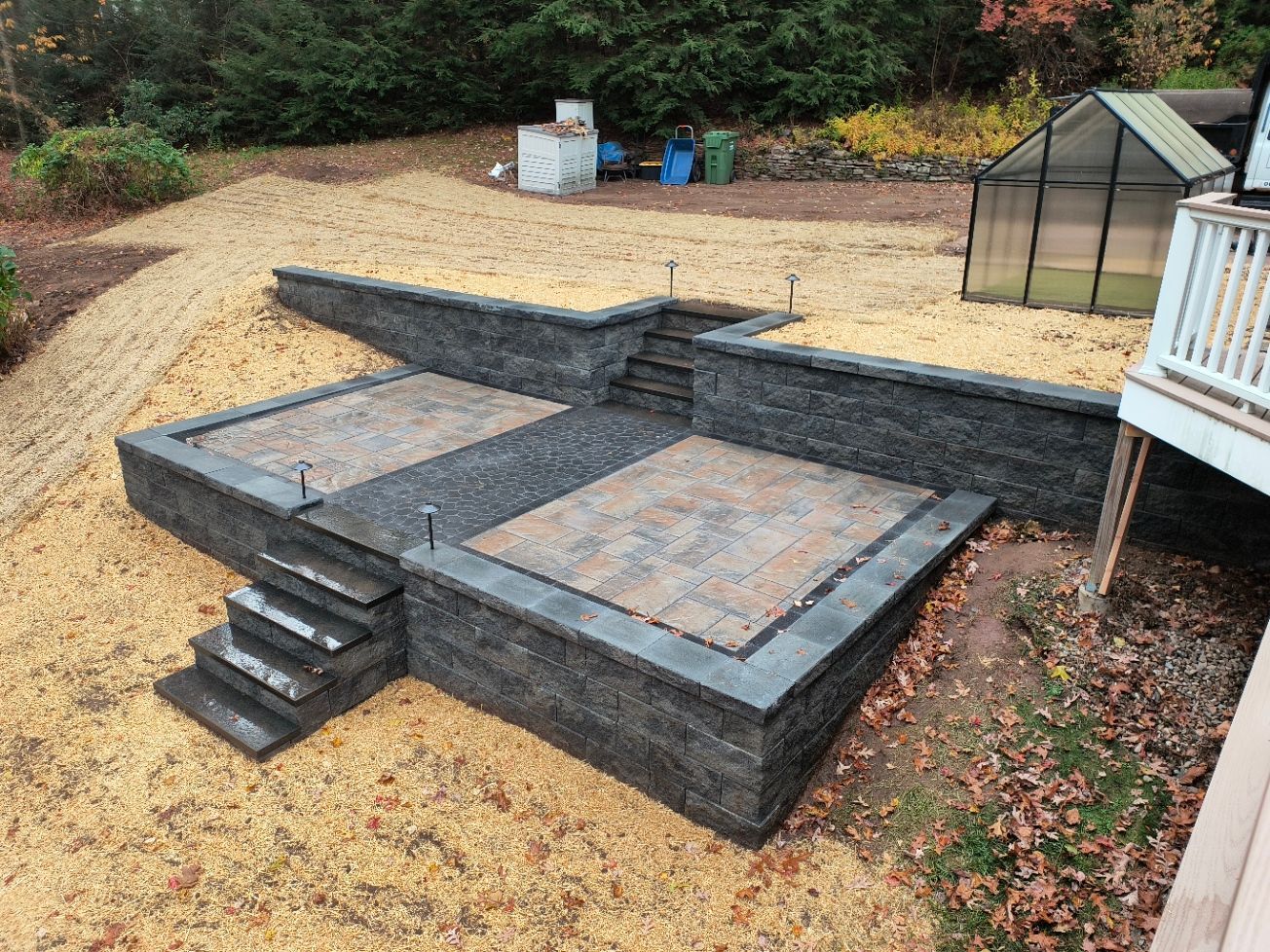 Stone patio with steps and retaining walls, surrounded by gravel and foliage. Greenhouse and deck visible.