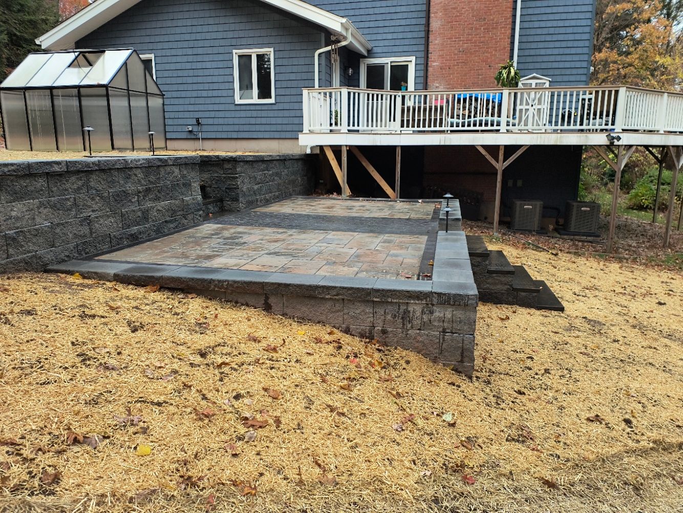 A house with a raised deck, a stone patio, and a greenhouse; yard covered in wood chips.