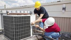 Two men are working on an air conditioner on the roof of a building.