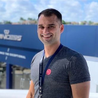 Smiling man in a dark gray shirt standing outdoors in front of a blue building.