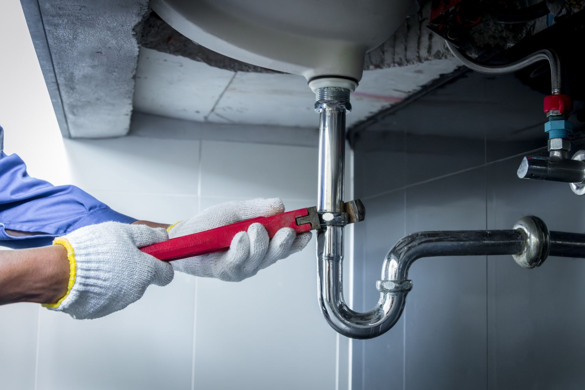 Plumber tightening a red pipe wrench on under-sink plumbing with metal pipes and a drain