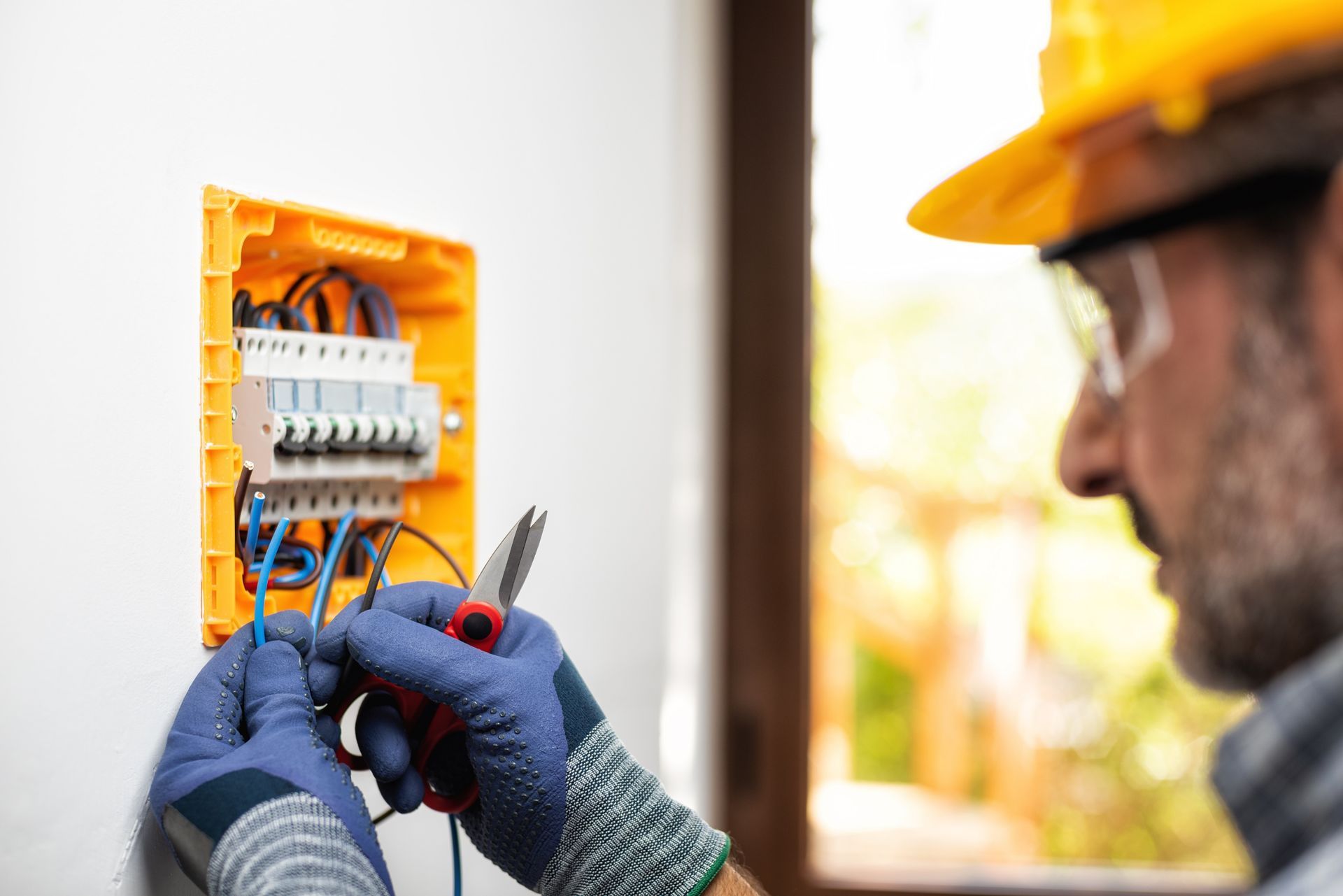 Electrician wiring a breaker panel in a wall, wearing gloves and a yellow hard hat.
