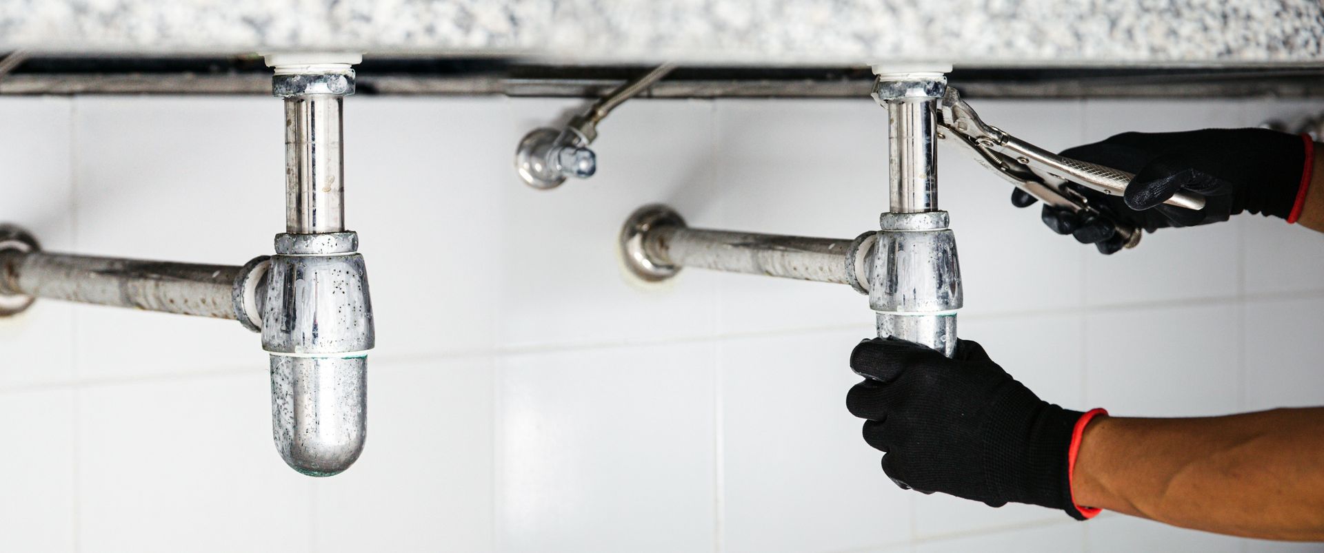 Plumber tightening a faucet under a sink with a wrench and black gloves.