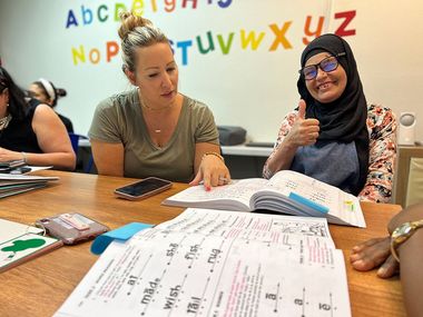 Two women in a classroom, studying open books. One gives a thumbs-up. A colorful alphabet is in the background.