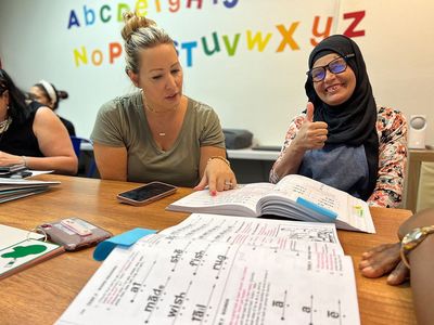 Two women in a classroom, studying open books. One gives a thumbs-up. A colorful alphabet is in the background.
