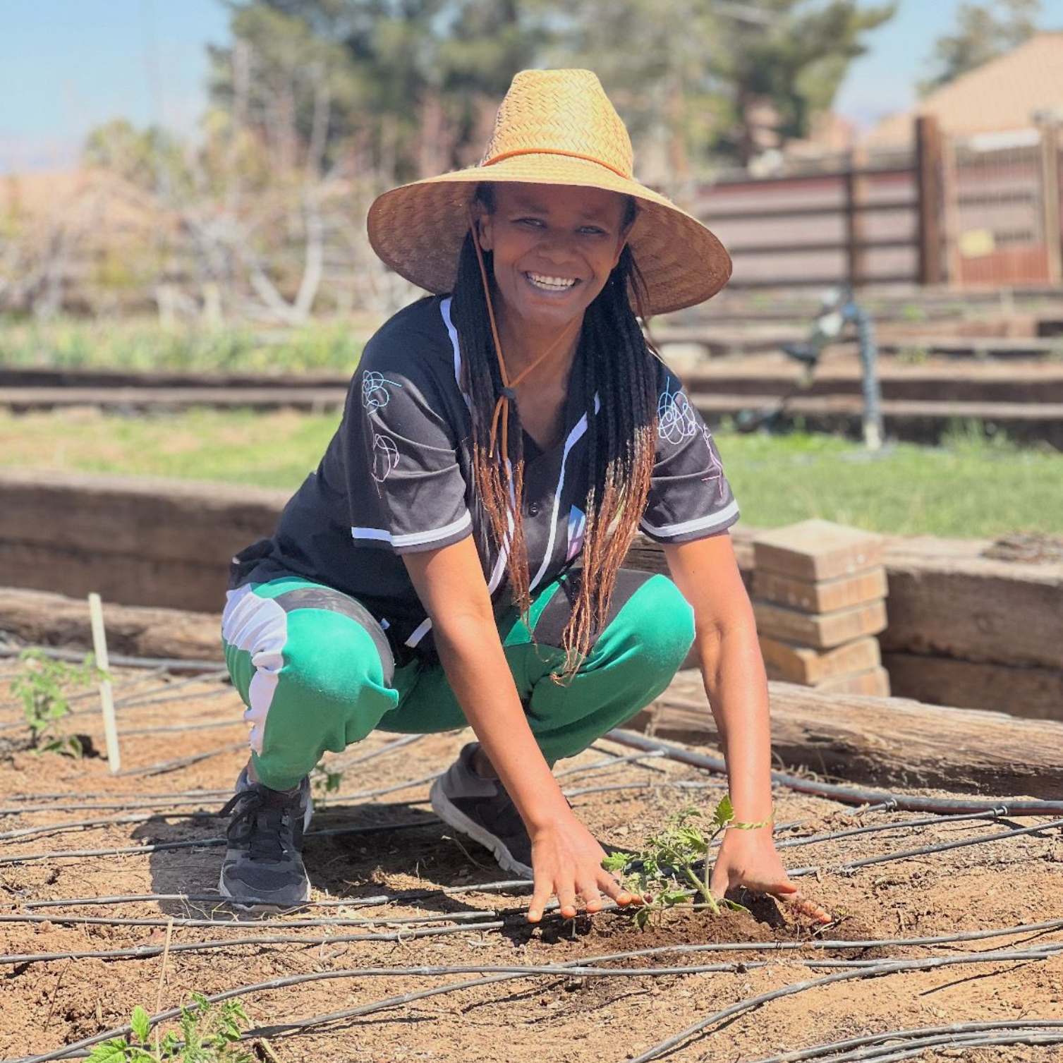 Woman wearing straw hat, kneeling in a garden bed, planting seedlings; smiling.