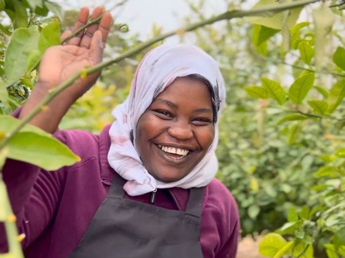 Woman in a hijab, apron, and purple shirt smiles amongst green plants, raising a hand.