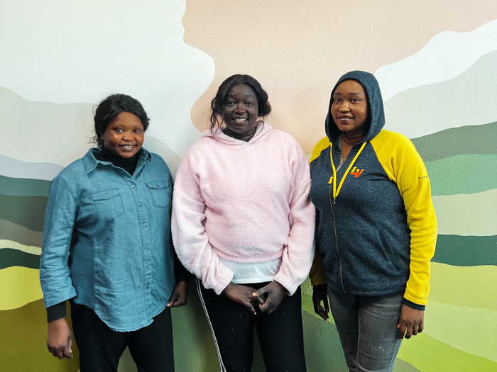 Three women smiling in front of a painted wall. One in blue denim, one in pink, one in a blue and yellow hoodie.