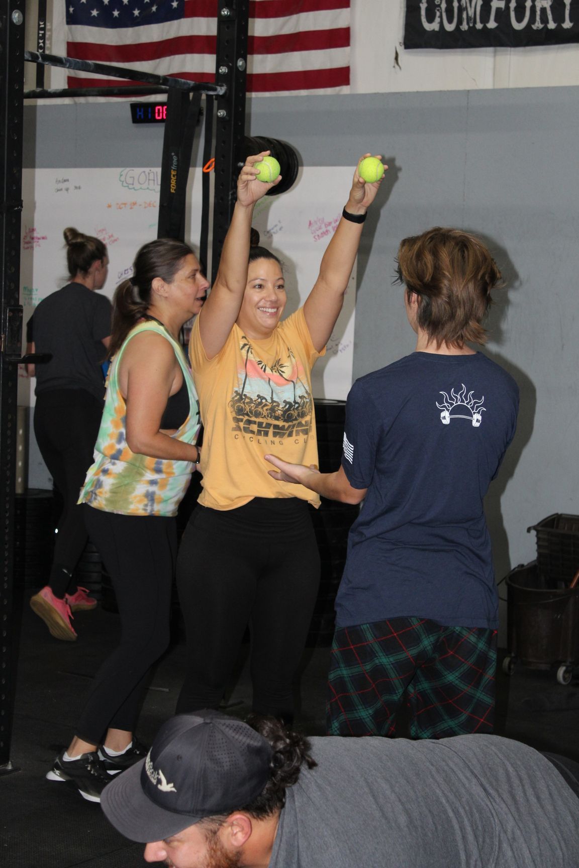 Three women in a gym, two lifting tennis balls overhead while another stands nearby.