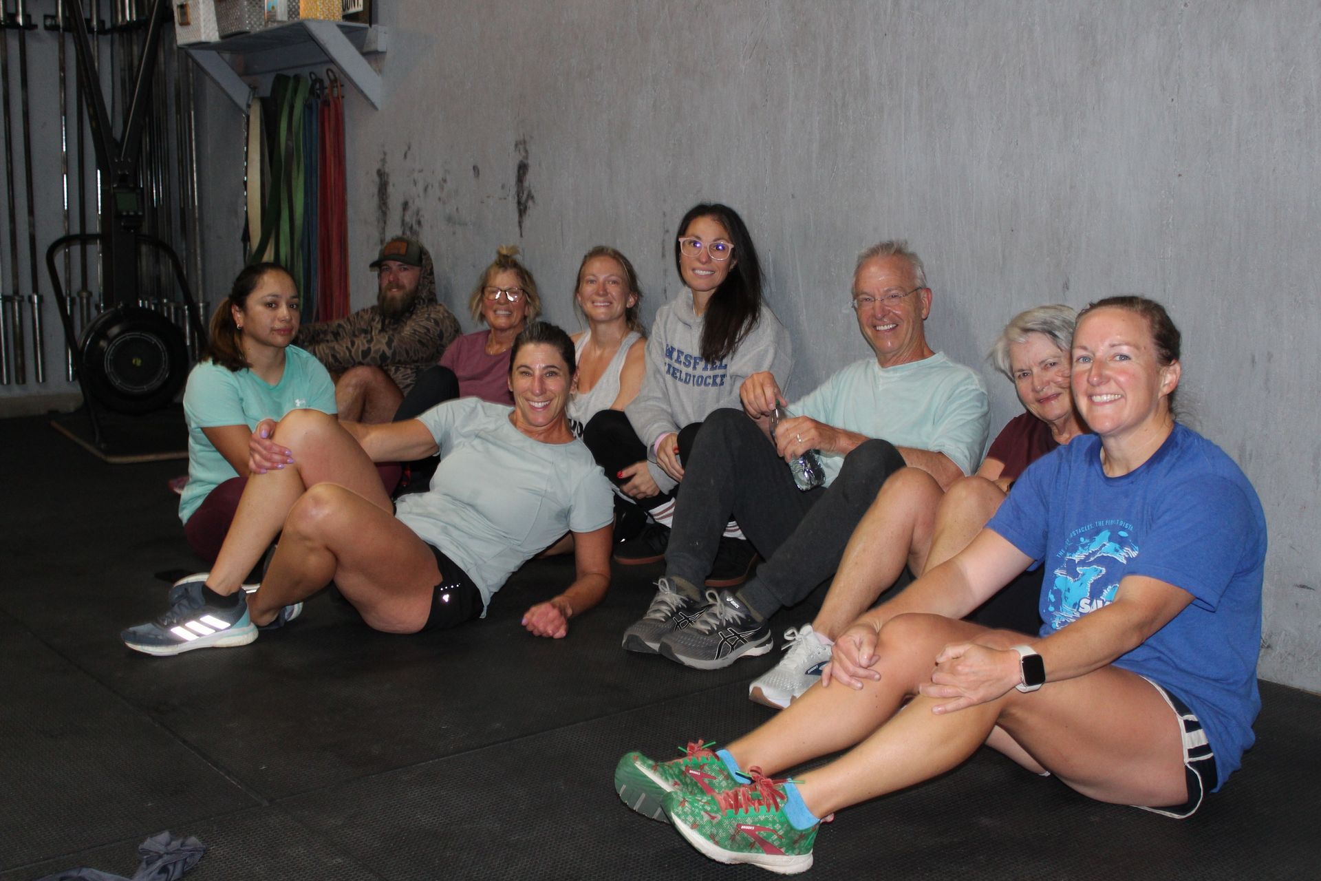 Group of people sitting on gym floor, smiling and posing in workout clothes.
