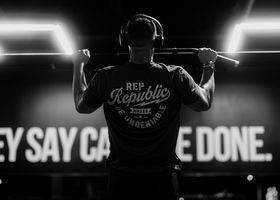 Backlit athlete lifting a barbell in a gym, with “Rep Republic” on shirt and motivational sign behind.