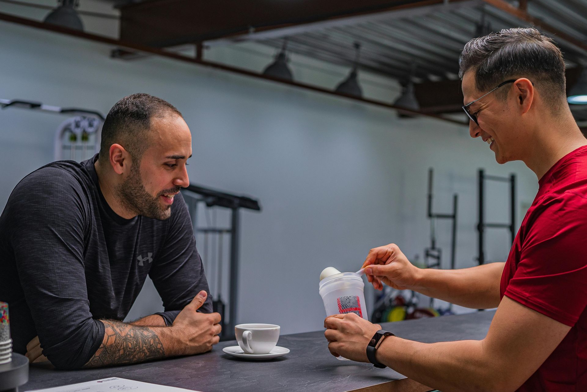Two men talking at a table, one holding a small item, in a gym or training space.