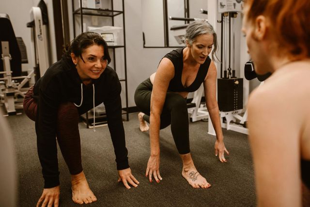 Two women in workout clothes stretching in a gym, with a trainer or classmate in the foreground.