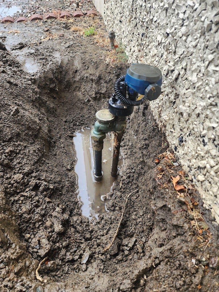 A water meter is sitting in the mud next to a wall.
