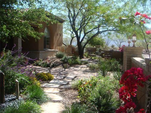 A stone walkway leading to a house surrounded by flowers and trees.