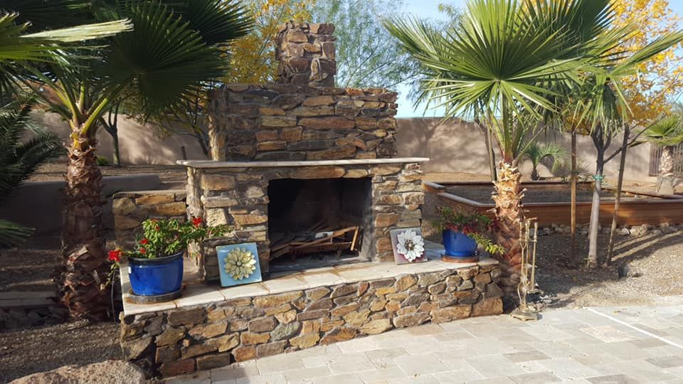 A stone fireplace is surrounded by palm trees on a patio.