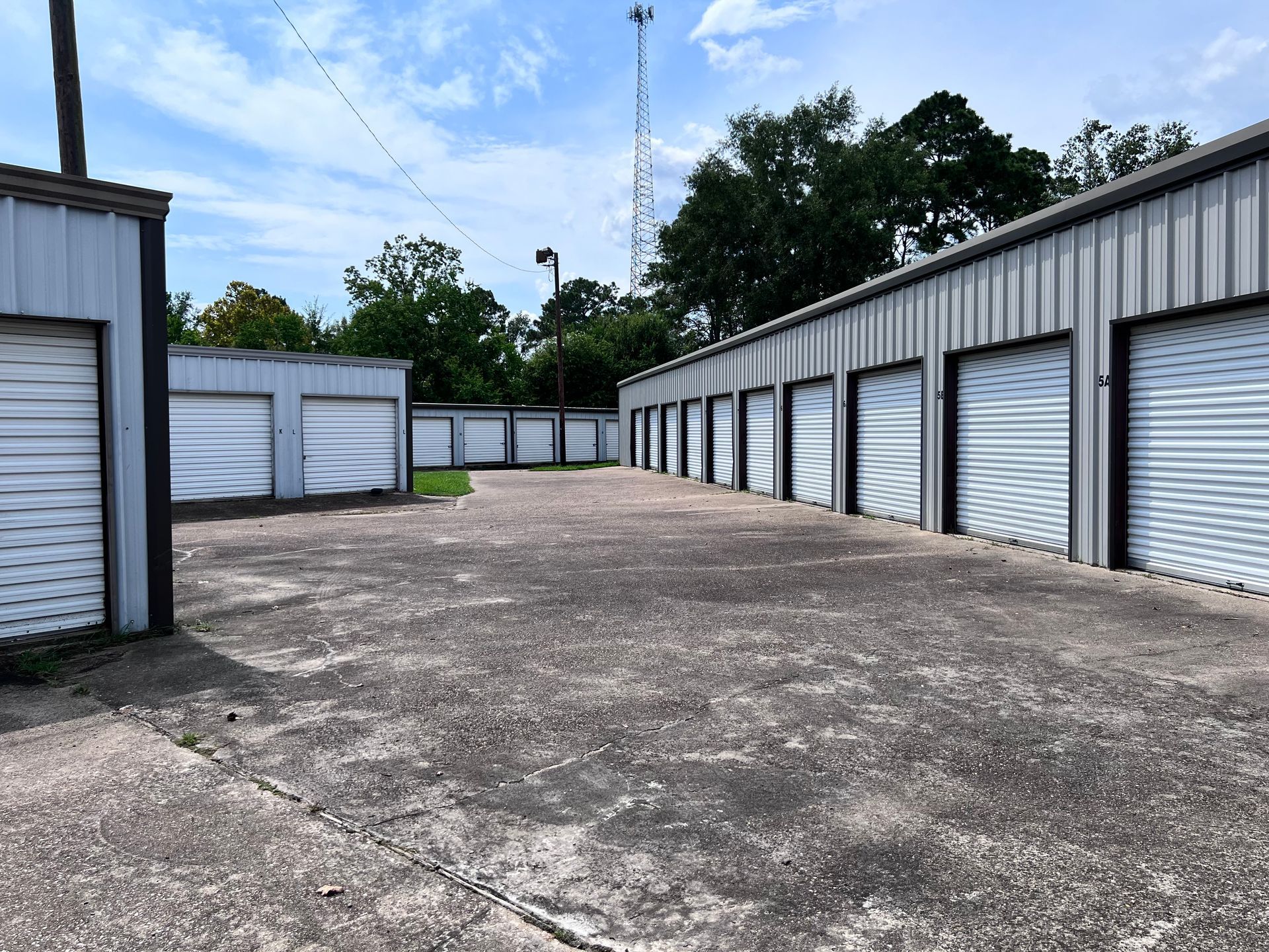 Storage units with gray doors, concrete ground, and a blue sky with trees in the background.