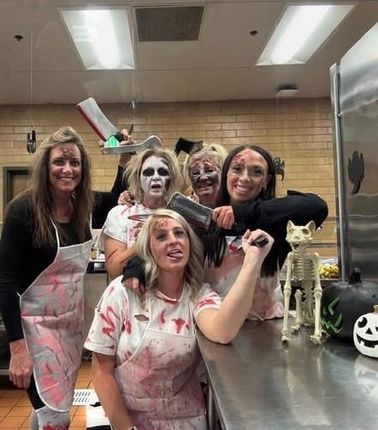Five people in zombie costumes in a kitchen. They wear blood-splattered aprons and hold props like cleavers and knives, Halloween decorations.