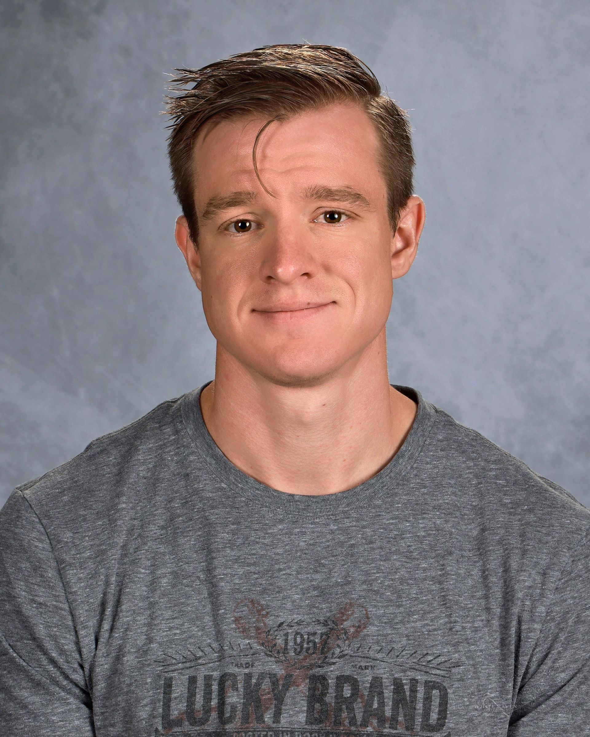 Man with fair skin and auburn hair smiles slightly. He wears a gray Lucky Brand shirt.