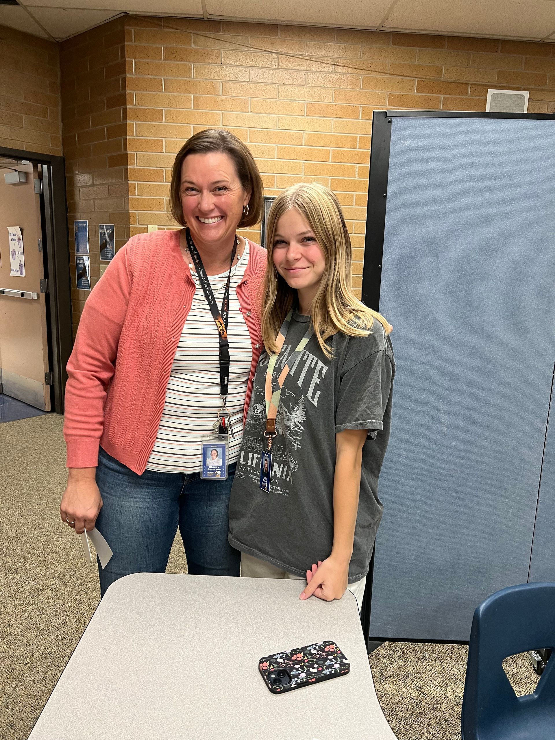 Woman in pink jacket and girl with long blonde hair smiling together by a desk in a classroom.
