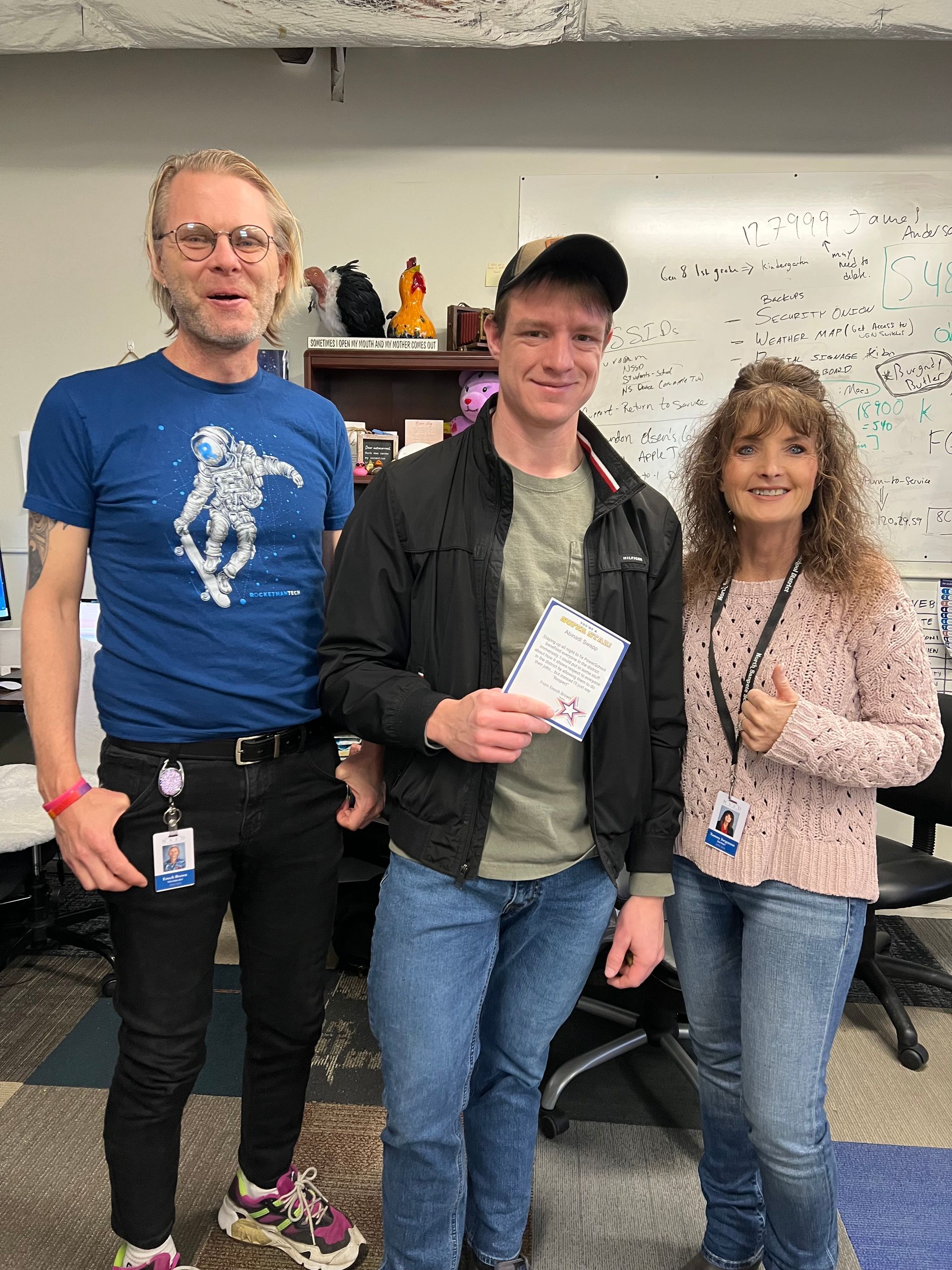 Three people smiling in an office. A man holds a paper, flanked by a man in a blue astronaut shirt and a woman in a pink sweater.