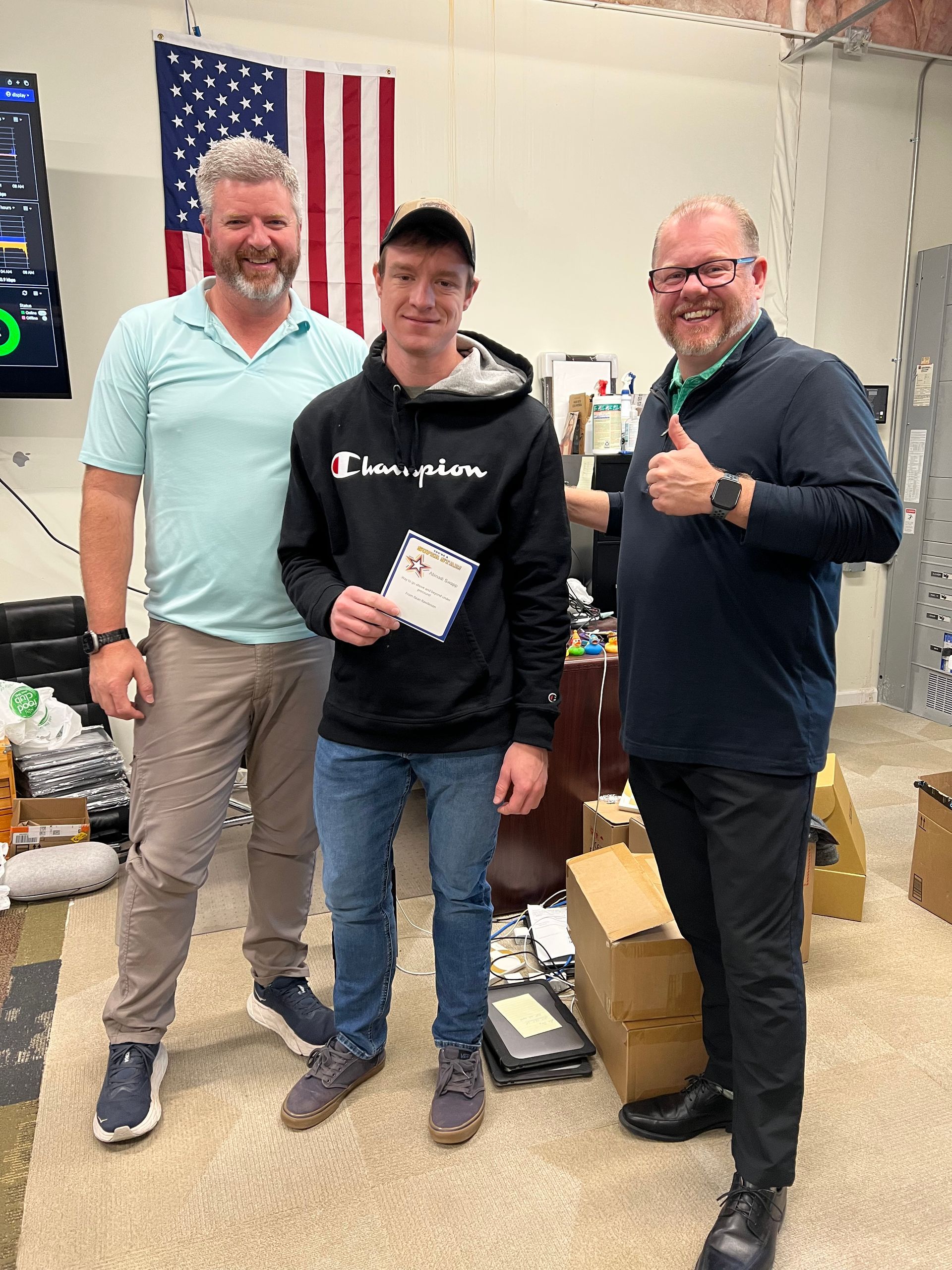 Three men pose indoors, one holding a paper. The man in the middle smiles. An American flag hangs on the wall.