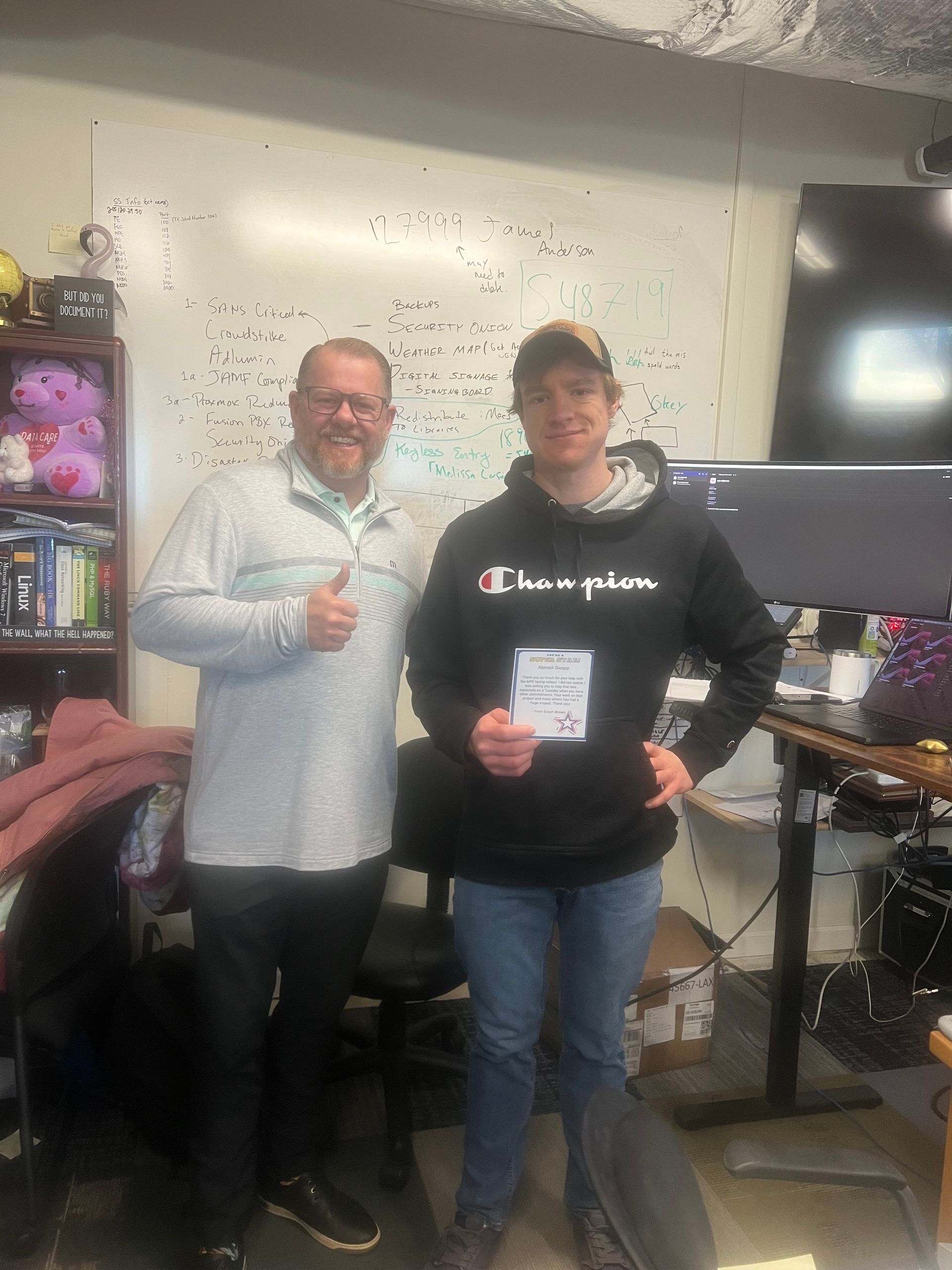 Two men pose together in an office. One gives a thumbs-up. The other holds a paper.