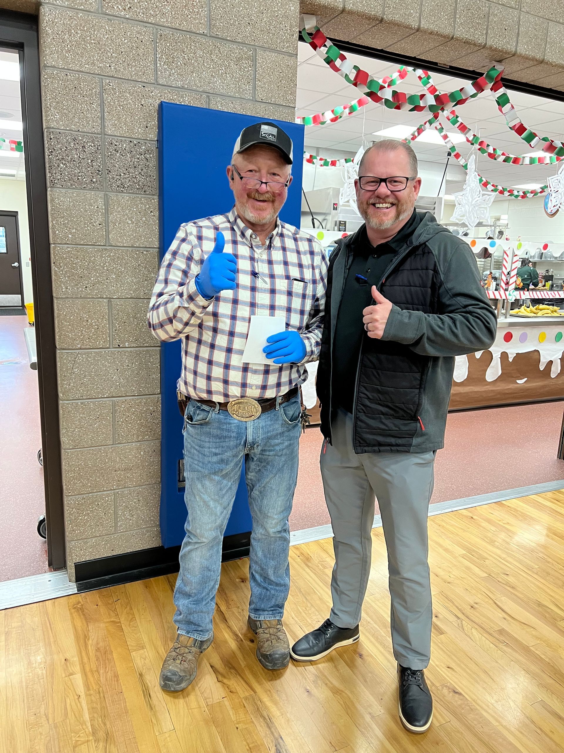 Two men giving thumbs up, smiling indoors. One in plaid shirt and jeans, other in a jacket and gray pants.