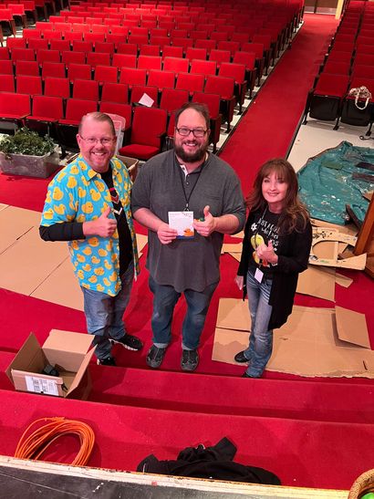 Three people are standing on a red carpet in an auditorium.