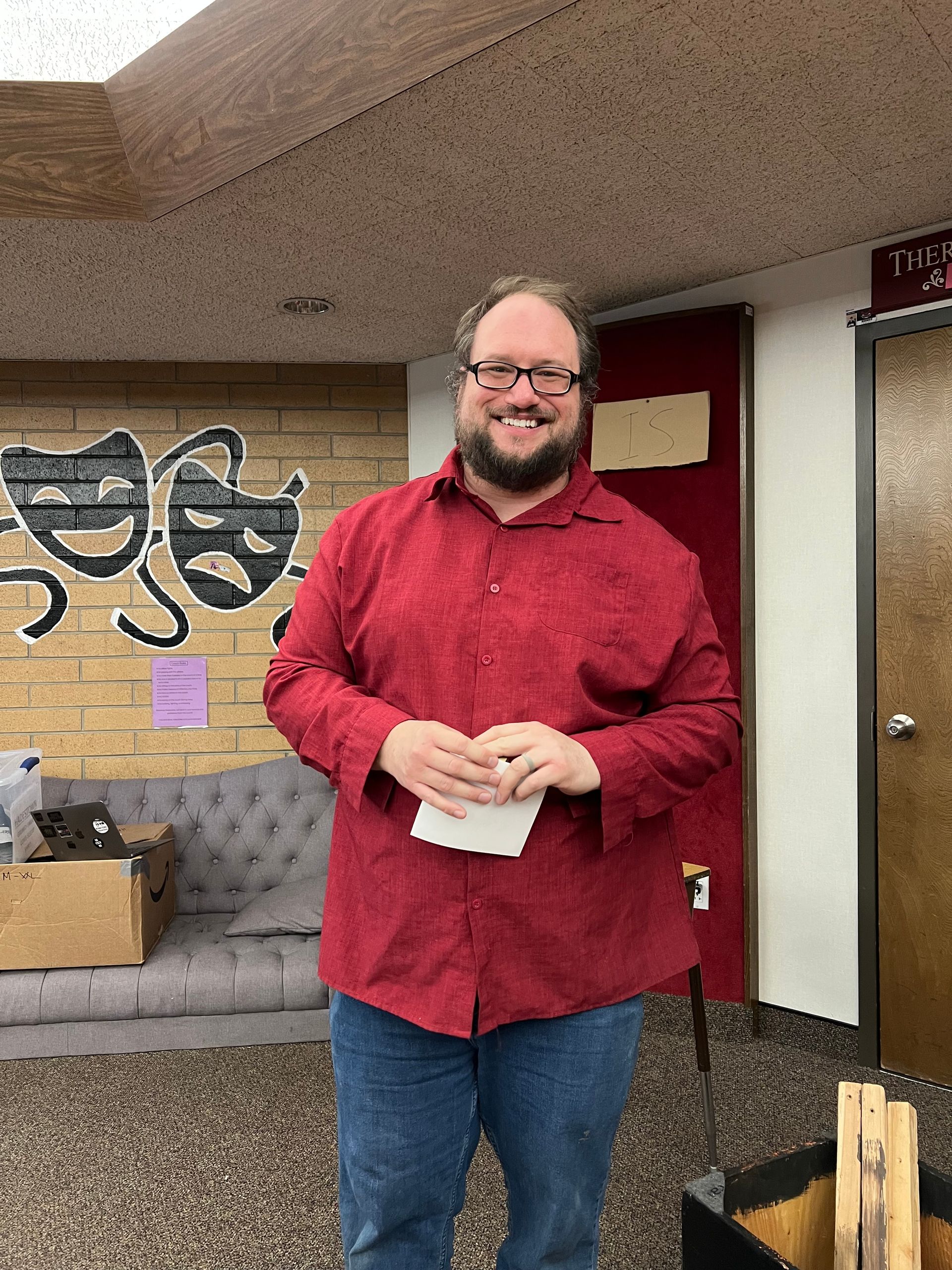 Smiling person in a red shirt stands indoors holding a paper, with a brick wall and office door behind them