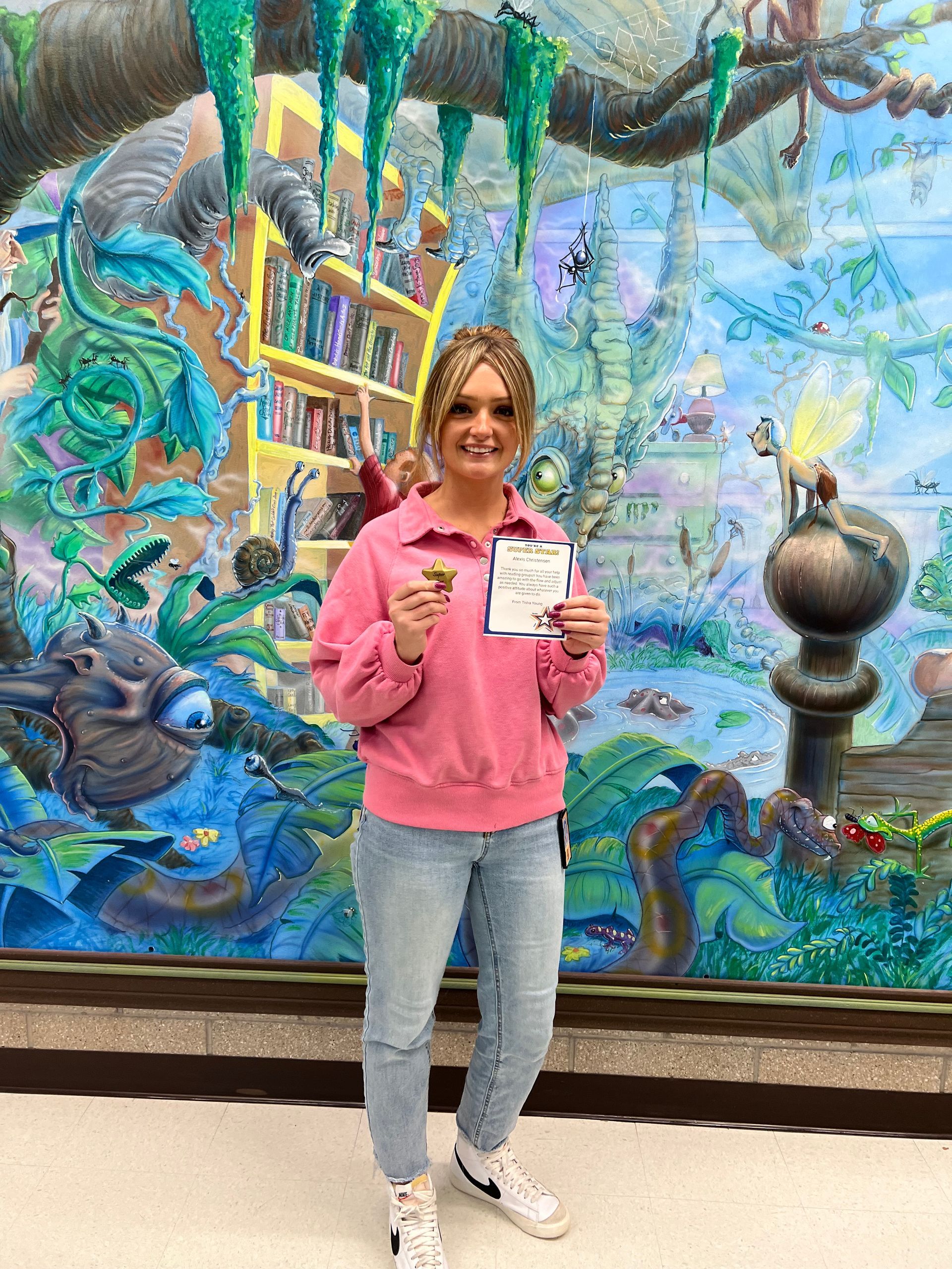 Woman in pink sweater and jeans stands in front of a mural, holding a certificate and object.