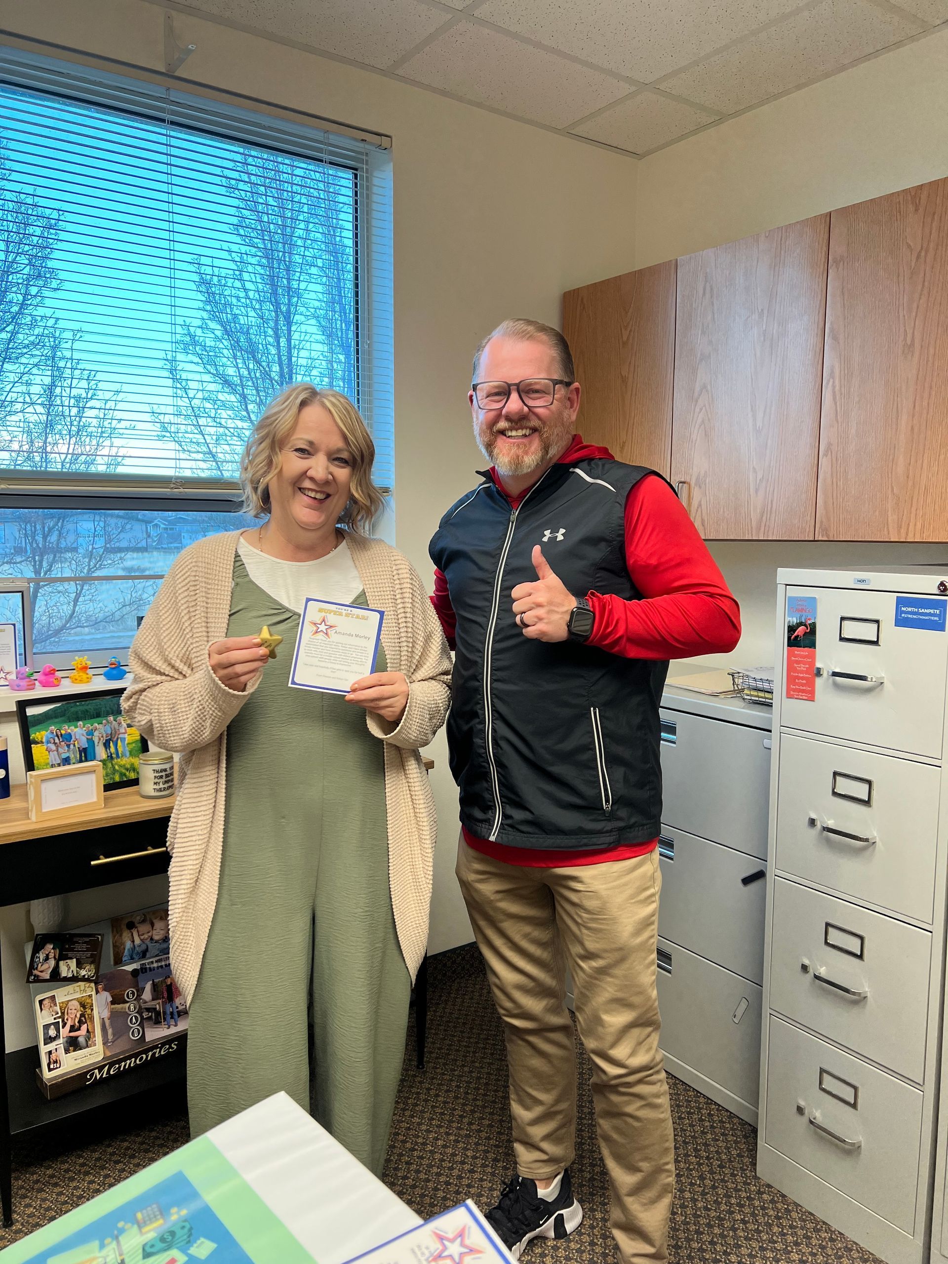 A smiling woman in a green jumpsuit holds a card next to a man giving a thumbs-up in an office with a window and cabinets.