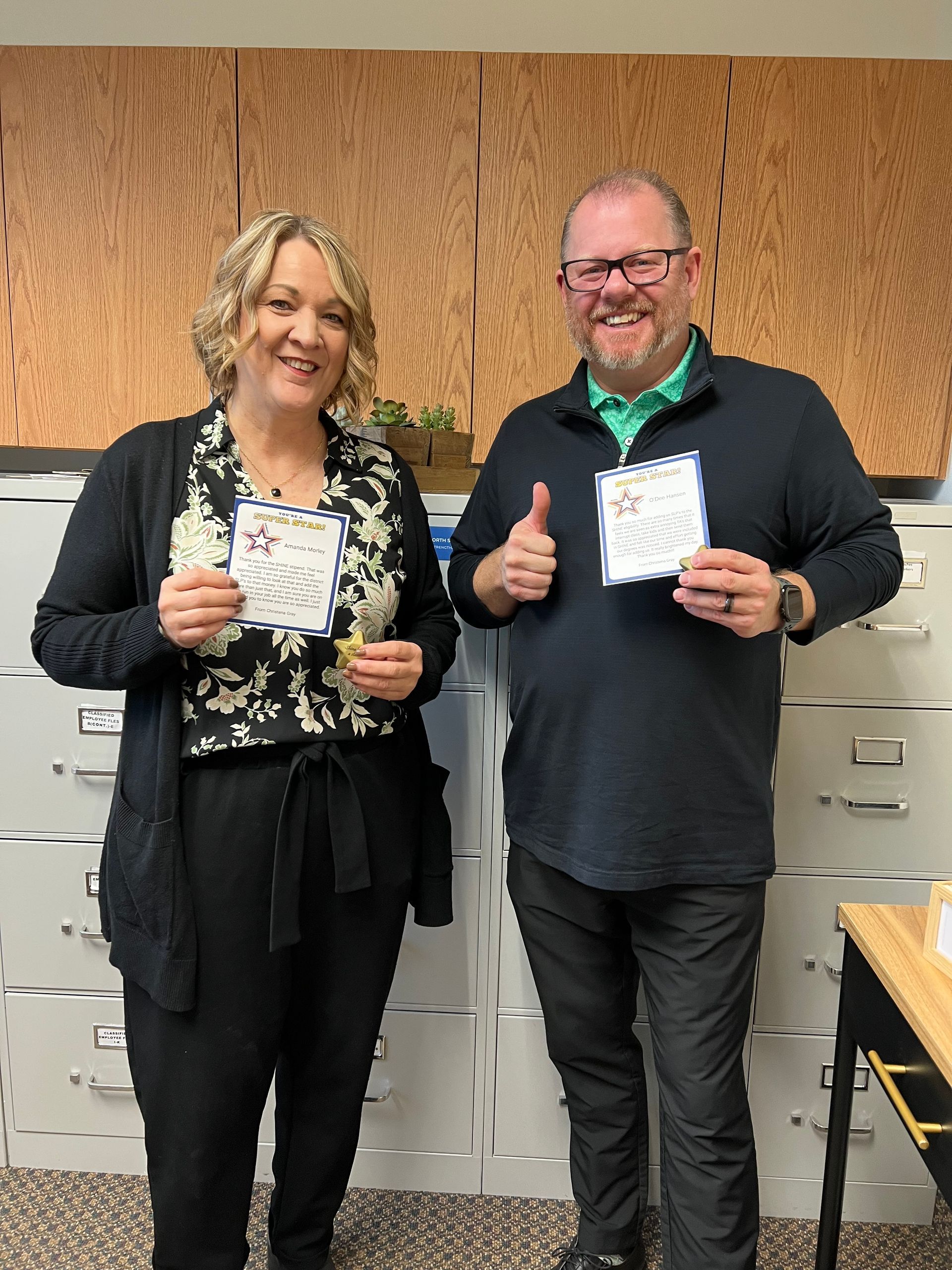 Two people holding certificates, smiling, standing in front of file cabinets and wood paneling.