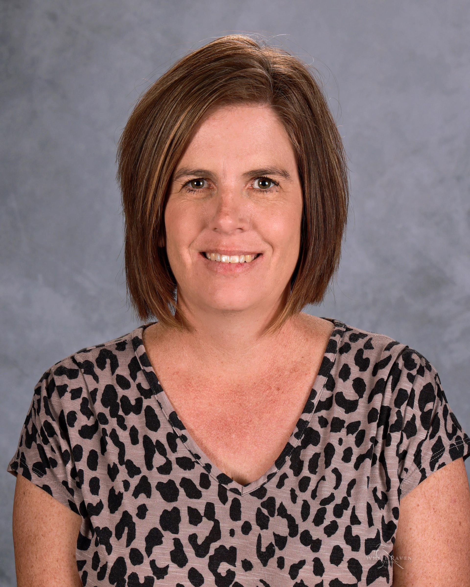 A person with shoulder-length brown hair, wearing a grey and black leopard-print top, smiles against a grey backdrop.
