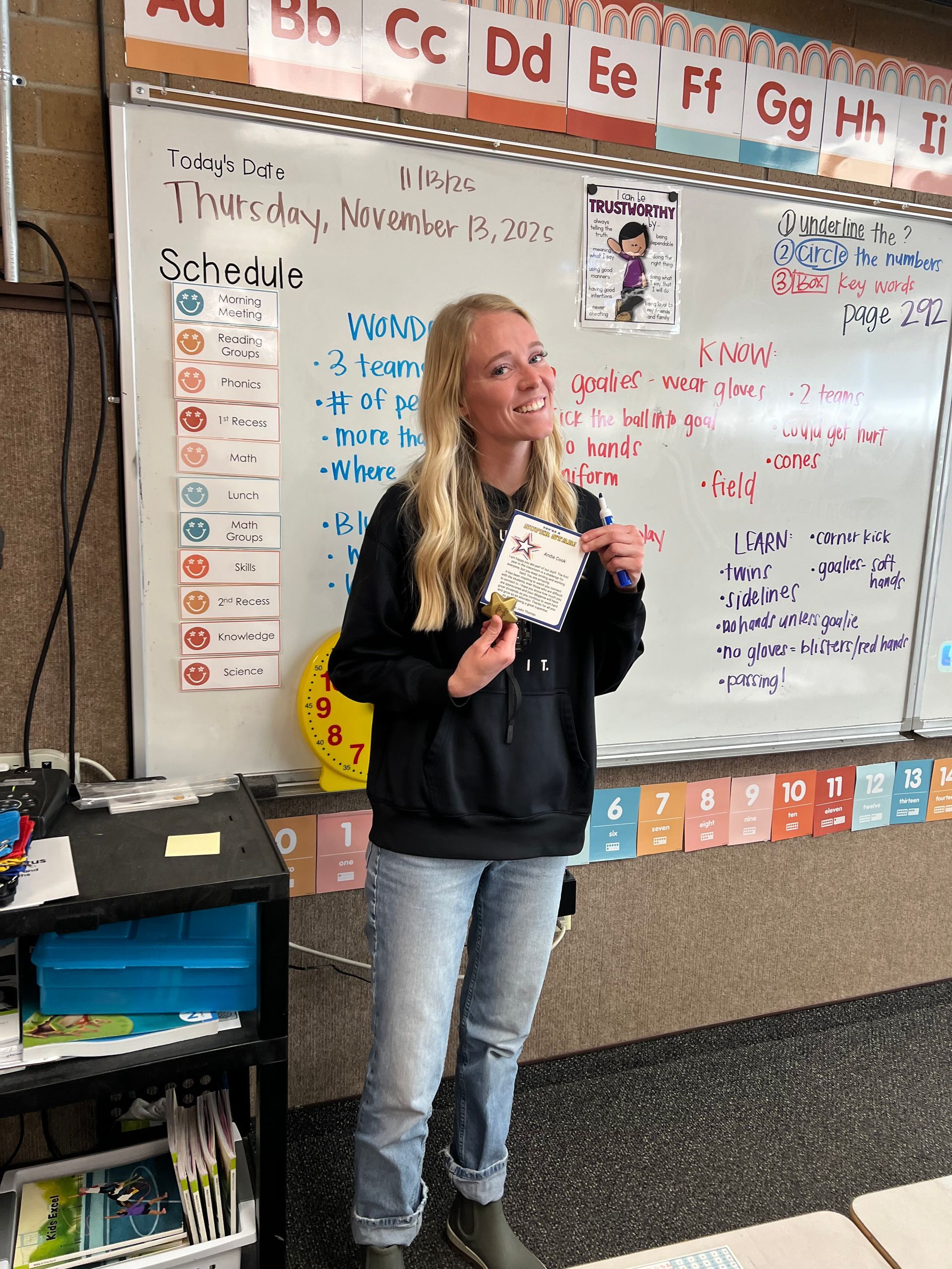 Teacher in black hoodie and jeans holds a paper in a classroom, standing in front of a whiteboard.