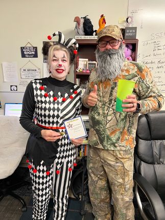 Two people in costume. One dressed as a jester, the other as a mountain man. Office setting.