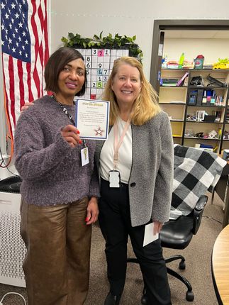 Two women pose in a classroom, holding a card. One woman wears a sweater, the other a blazer. American flag visible.