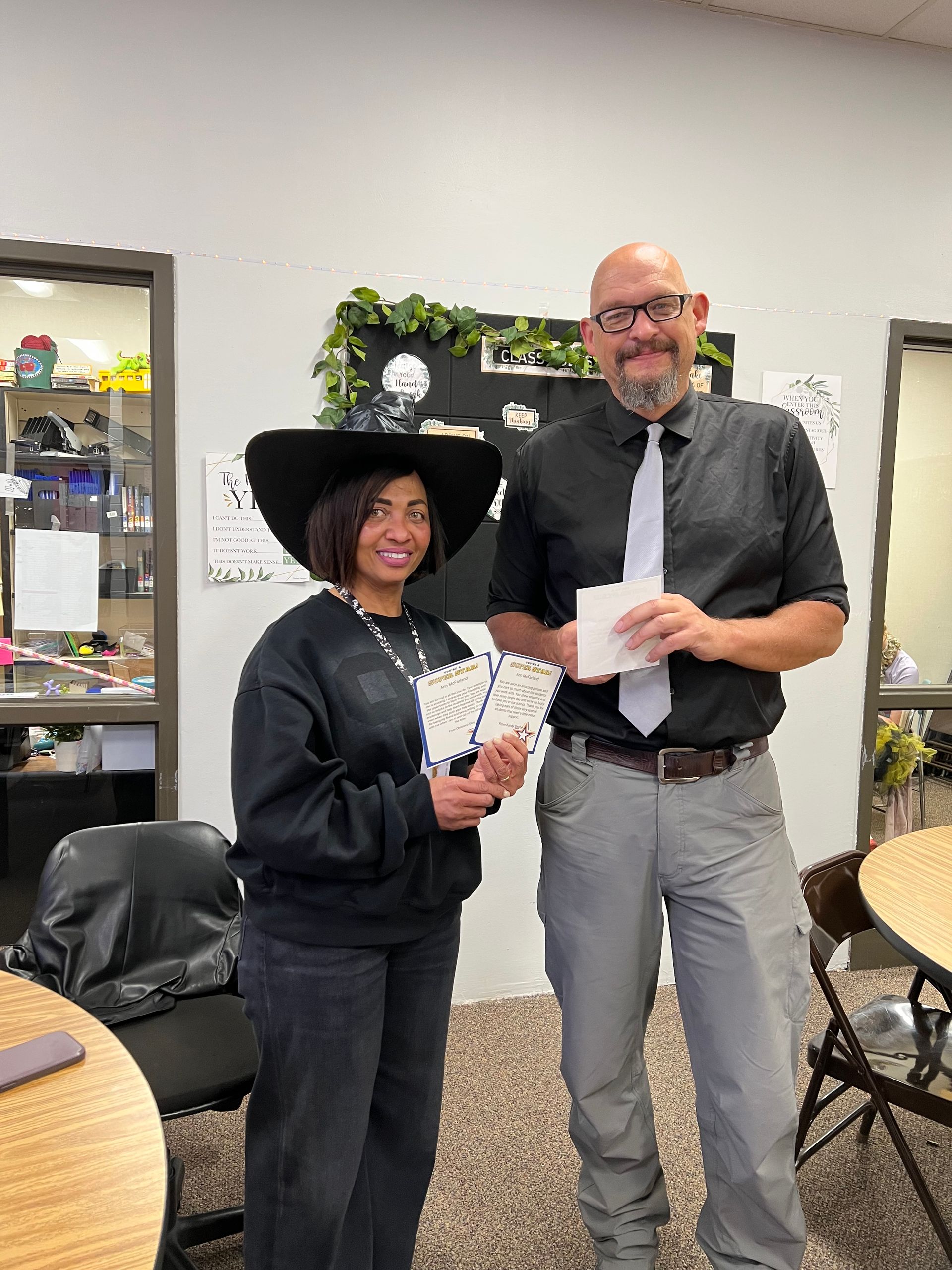A person in a witch hat and a man holding cards, standing indoors.