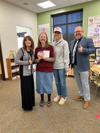 Four people pose indoors, smiling. A woman in a red shirt holds a paper. Two give thumbs up. Bright room.