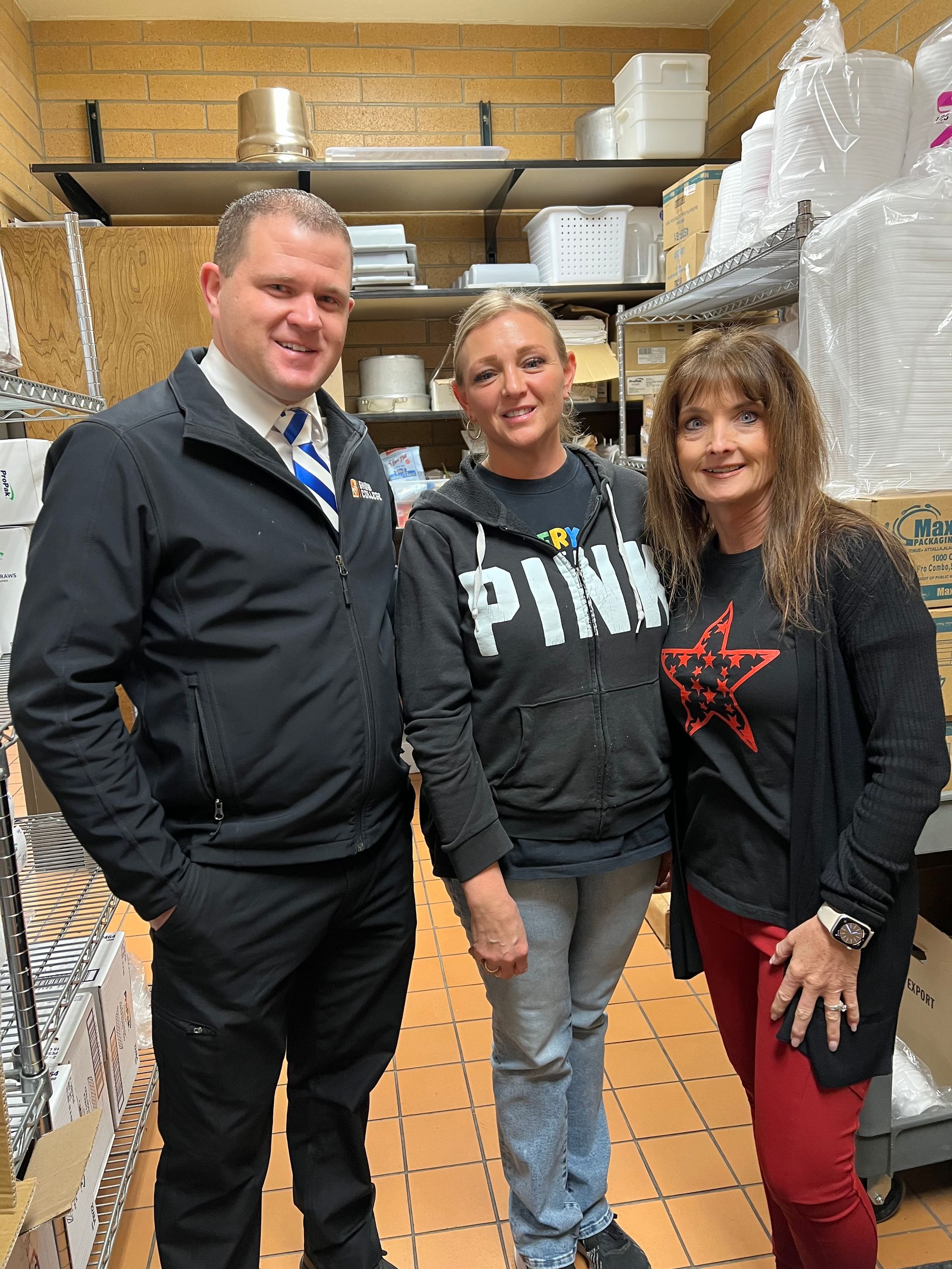 Three people smiling in a restaurant's storage area; man in suit jacket, two women in casual attire.