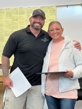 Two smiling people stand together indoors, holding papers and a folder, with a wall of notes behind them.