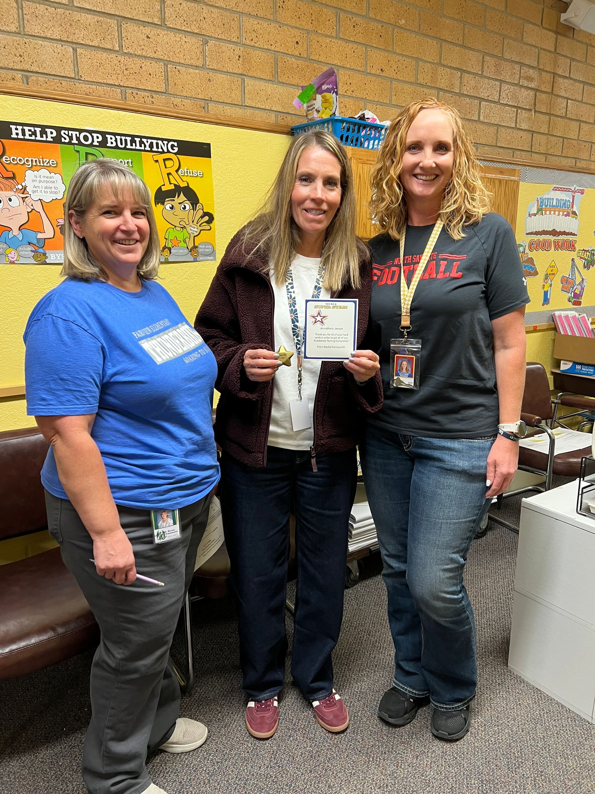 Three women standing together in a classroom; one holds an award.