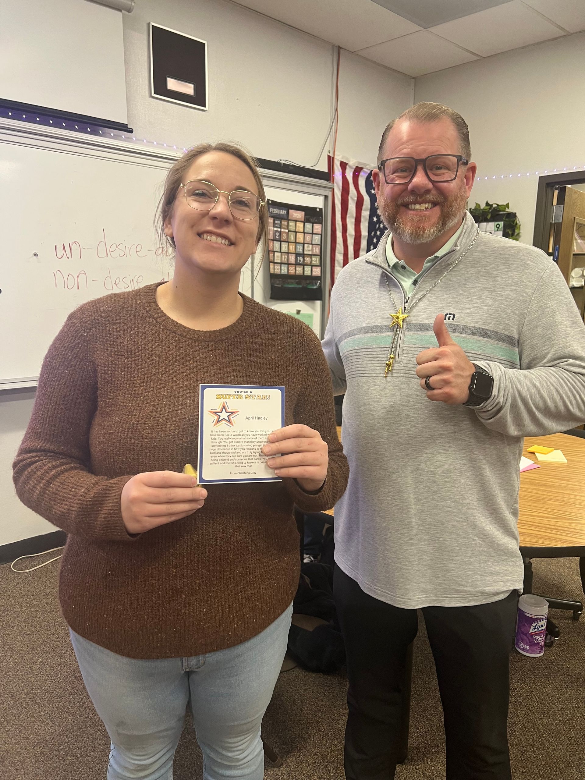 Woman holding a certificate smiles with a man giving a thumbs up in a classroom setting.
