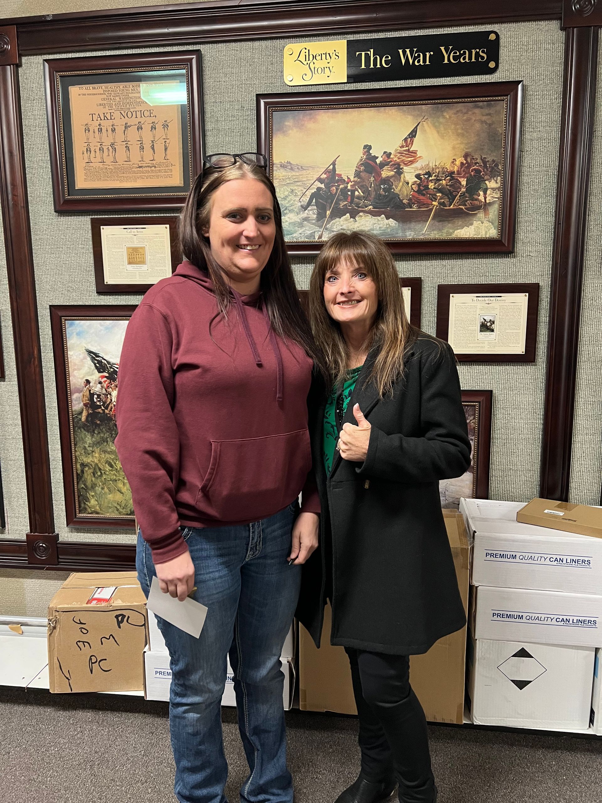 Two women pose next to boxes. One in a red hoodie and jeans, the other in a black coat giving a thumbs up. A wall behind them has framed historical documents and art.