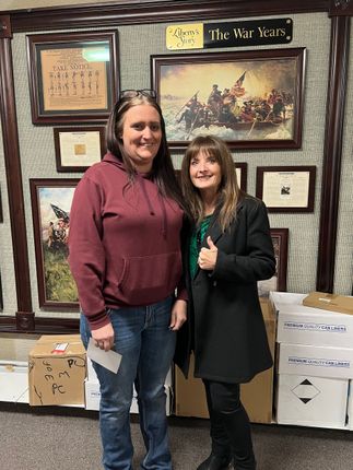 Two women smiling, posing in front of framed artwork. One in a red hoodie and jeans, the other in a black coat with a thumbs up. Boxes are in front of them.