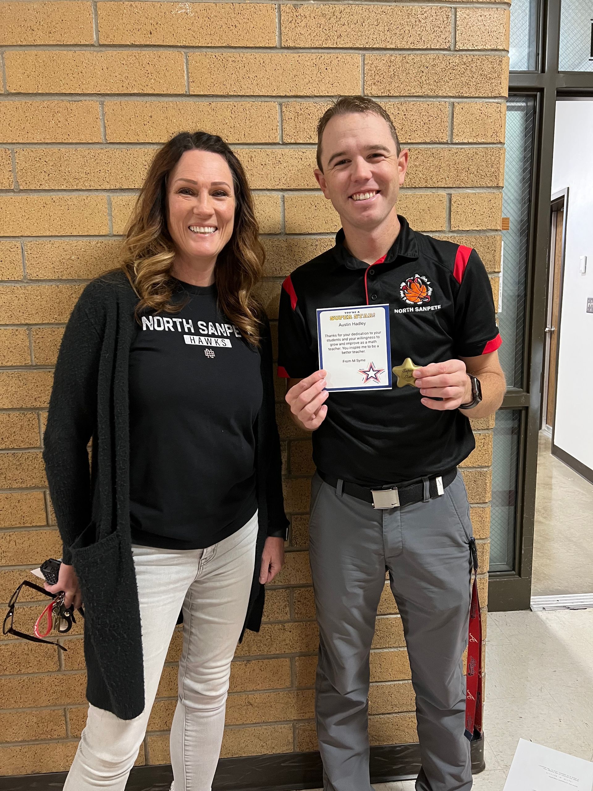 Woman and man smiling, posing near a brick wall; man holds a certificate and a small trophy.