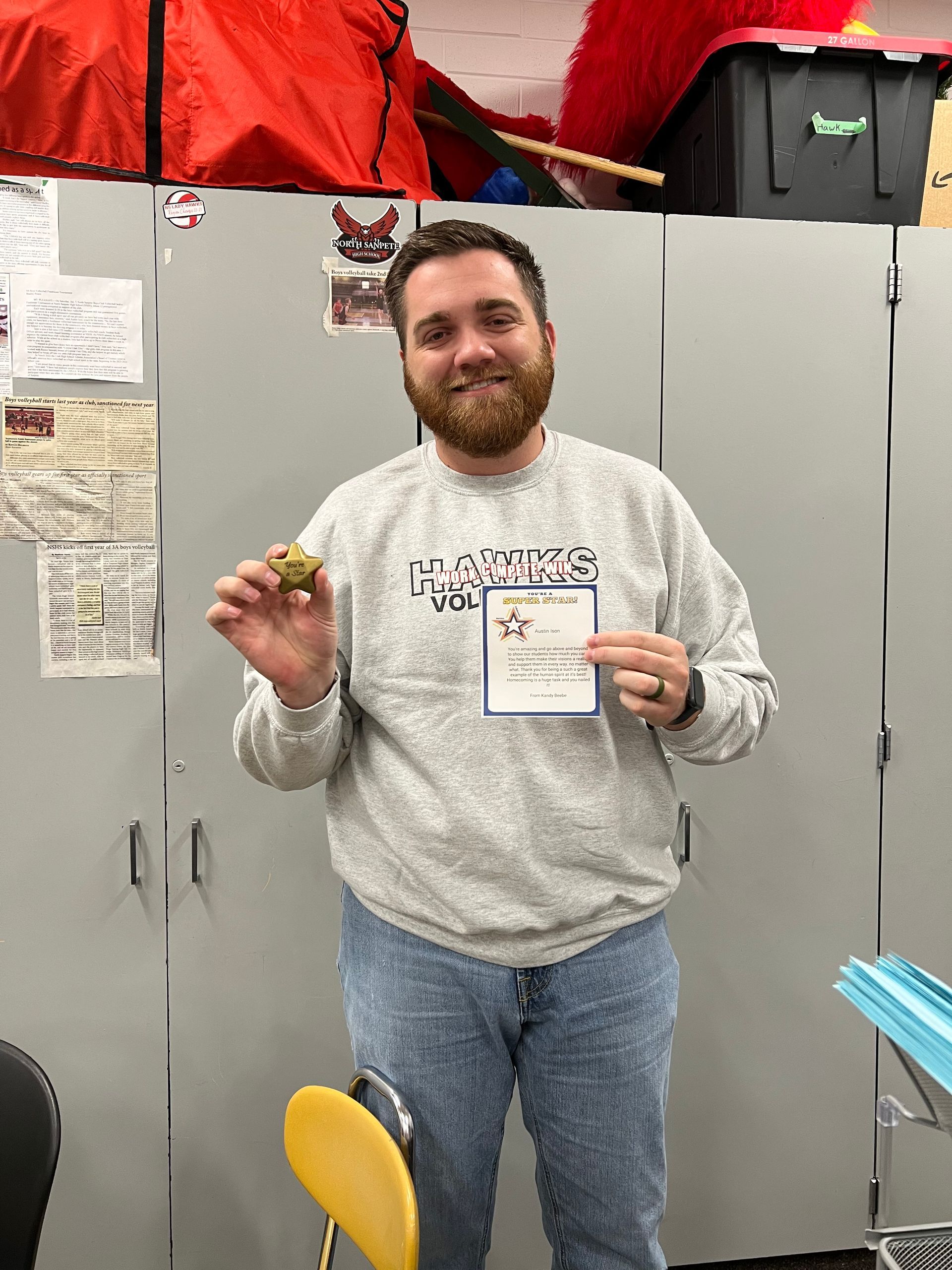 Man in gray sweatshirt and jeans holding a certificate and a small object, standing in front of gray lockers.