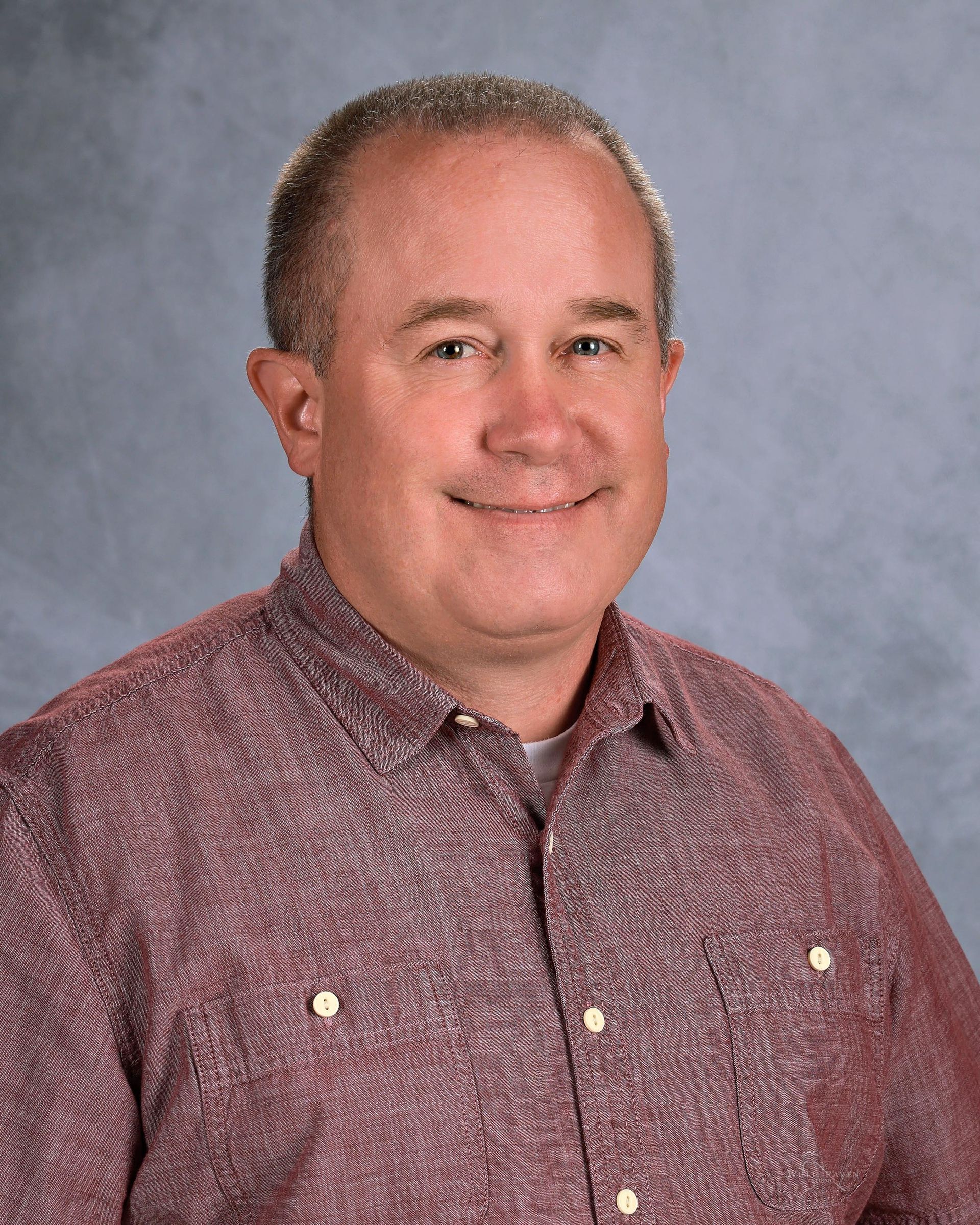 Middle-aged man with fair skin, smiling, wearing a red button-down shirt, against a gray backdrop.