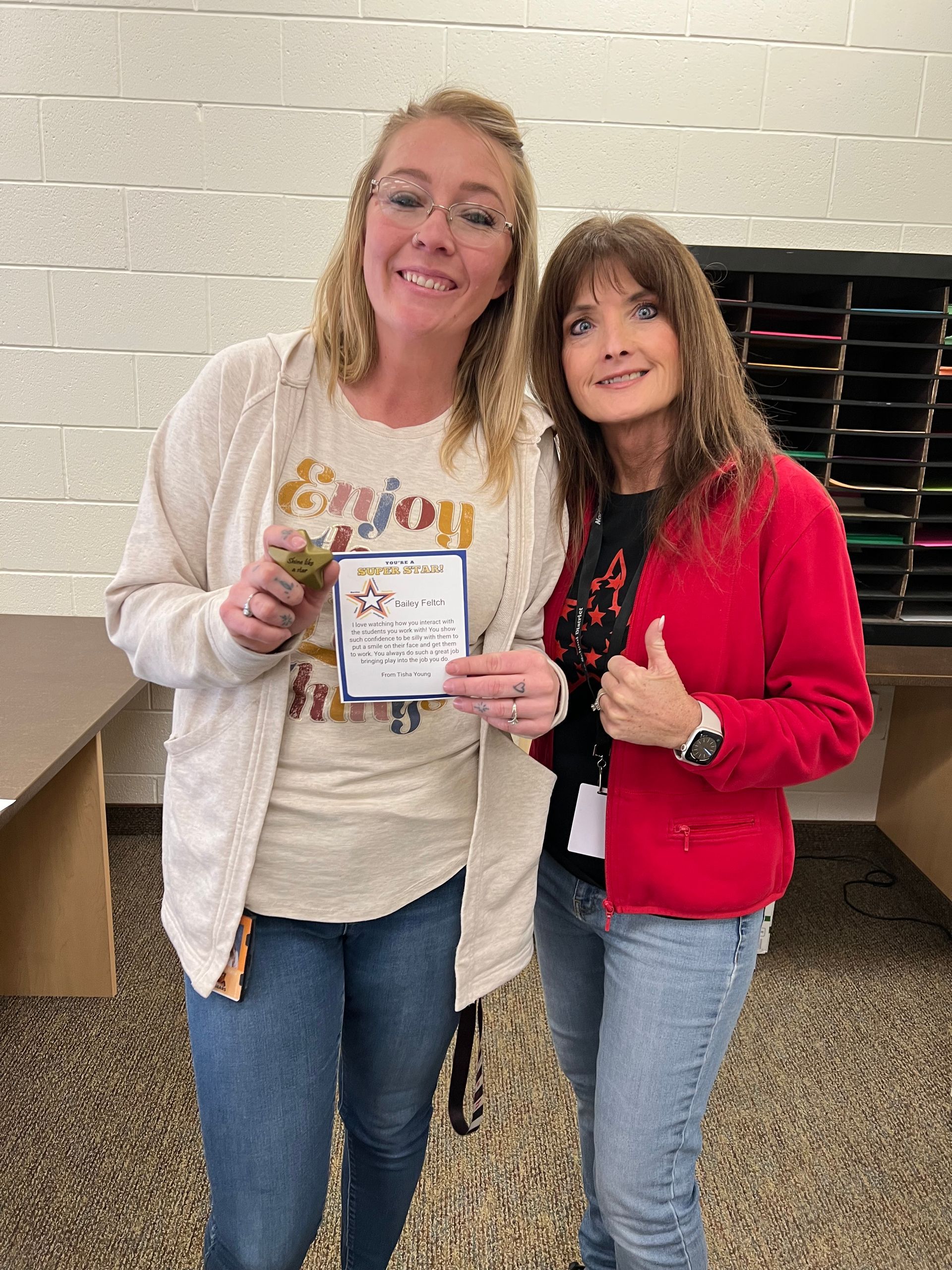 Two women smiling, one holding a gift and card. The other gives a thumbs up. Indoor setting.